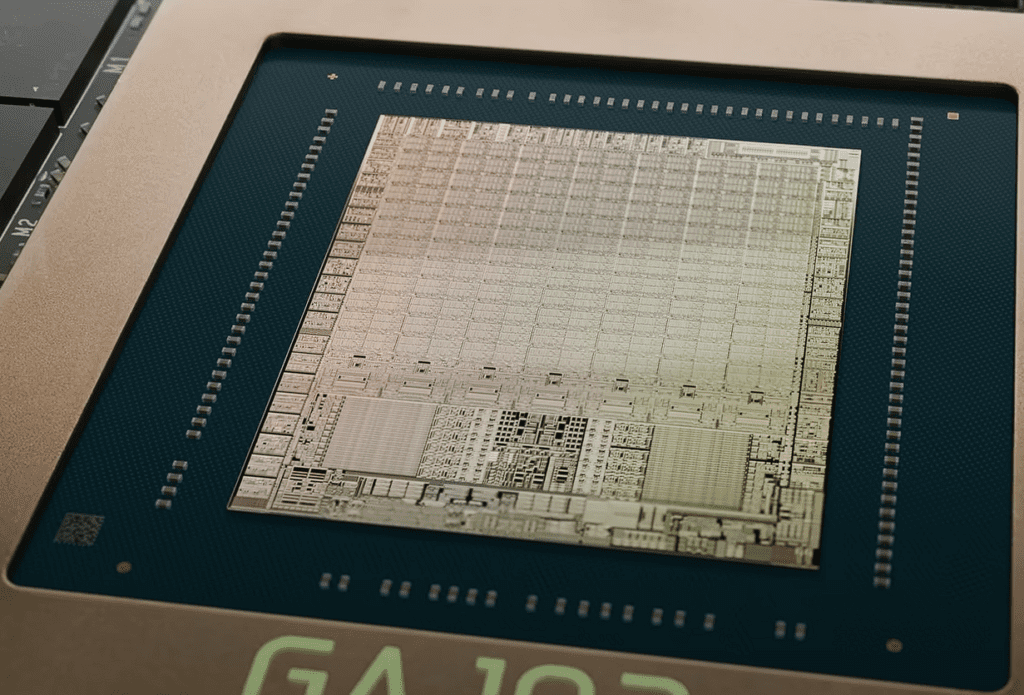A macro photograph of an exposed GPU silicon die (NVIDIA GA102) mounted on its substrate. The central square die displays a grid of circuit patterns and metal interconnects in gold and silver hues, with fine geometric blocks representing transistor arrays. Around the die’s perimeter are tiny bond pads and solder bumps, all seated within a teal-green package frame.